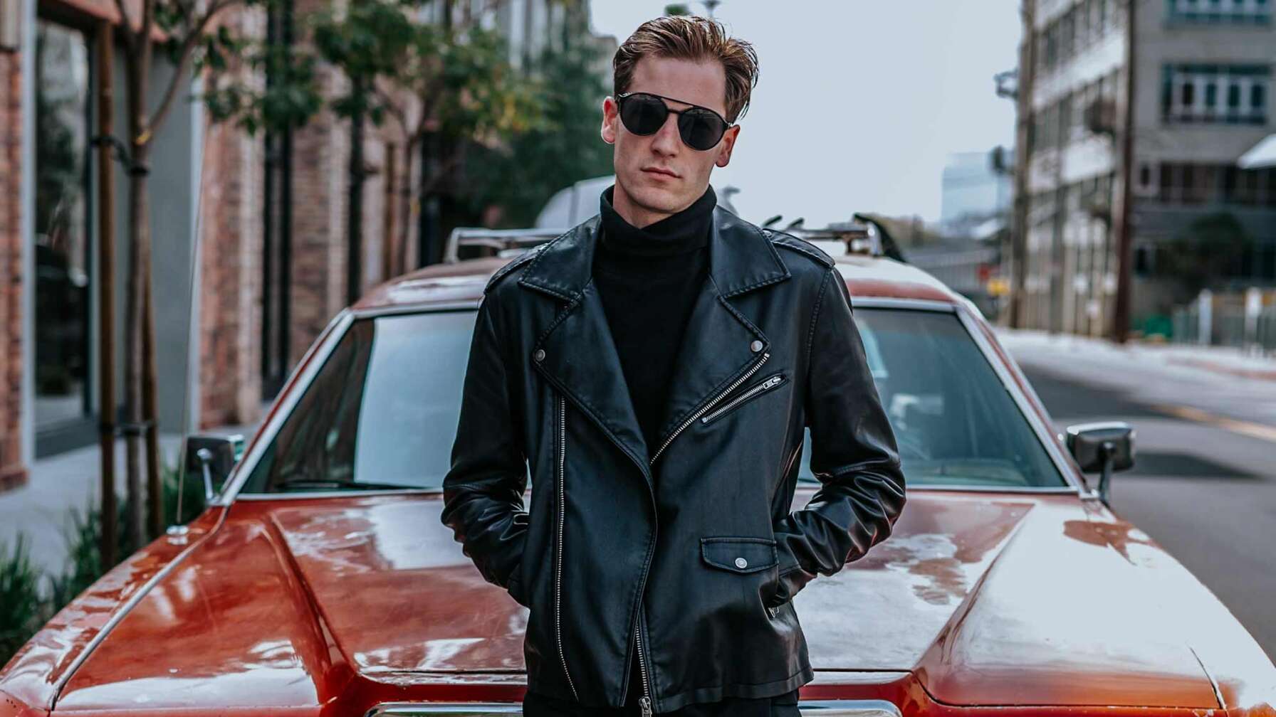 Bild eines jungen Mannes mit Lederjacke und Sonnenbrille, der an der Motorhaube eines roten Oldtimers lehnt